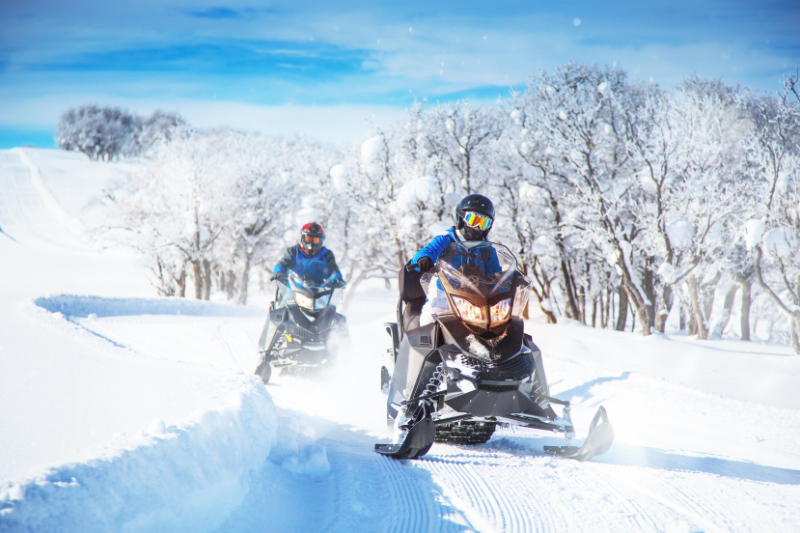 Two people riding snowmobiles wearing winter gear on a clear day